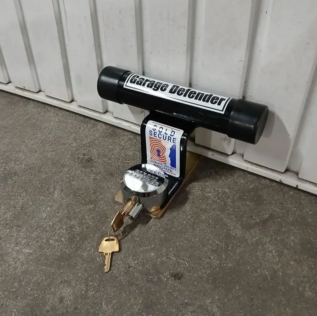 A black Garage Defender locked to a plate on a concrete floor against the bottom of a white ribbed garage door, keys in the padlock