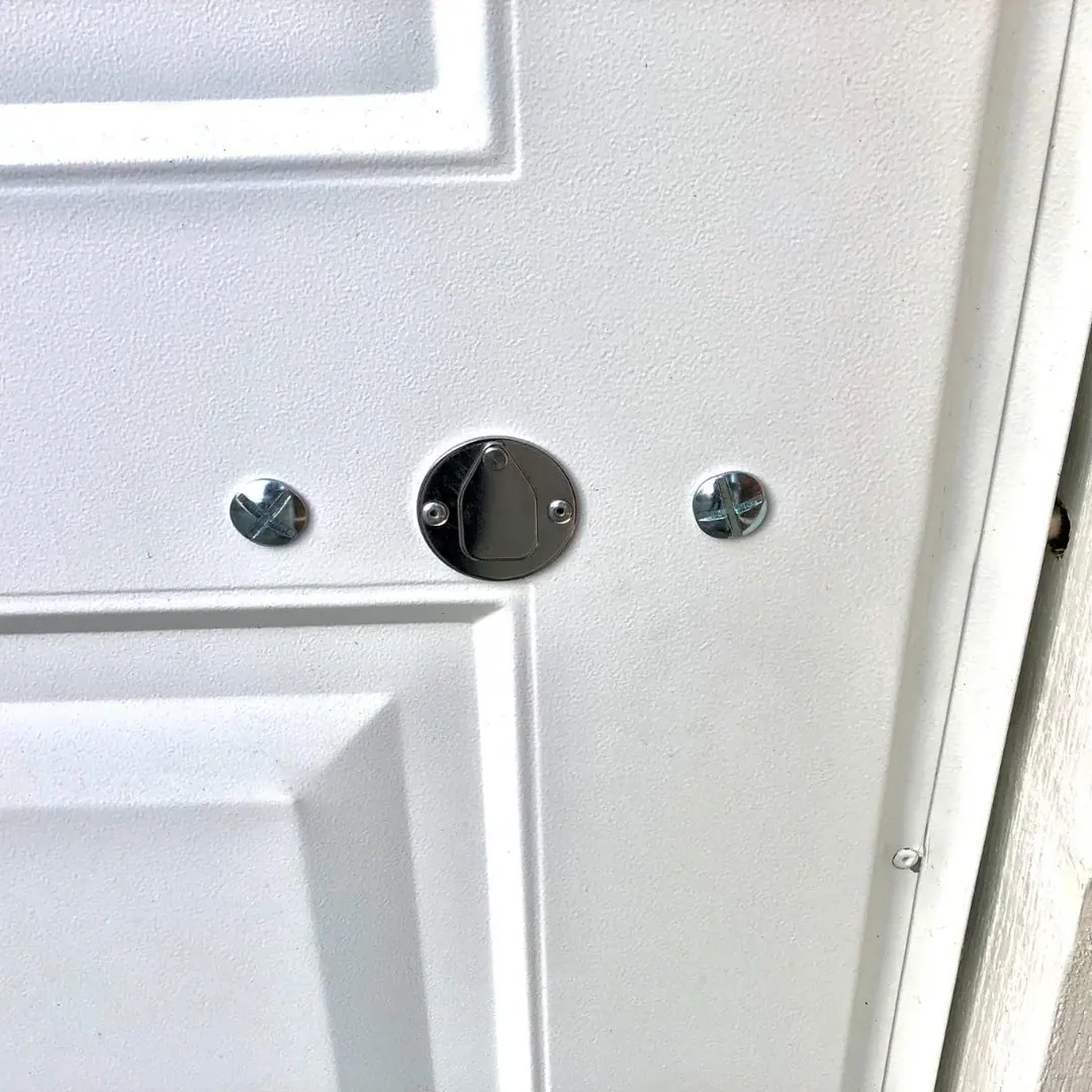 Close up of a white steel garage door with a round chrome keyhole cover and a hinged flap between two cross-head bolt heads