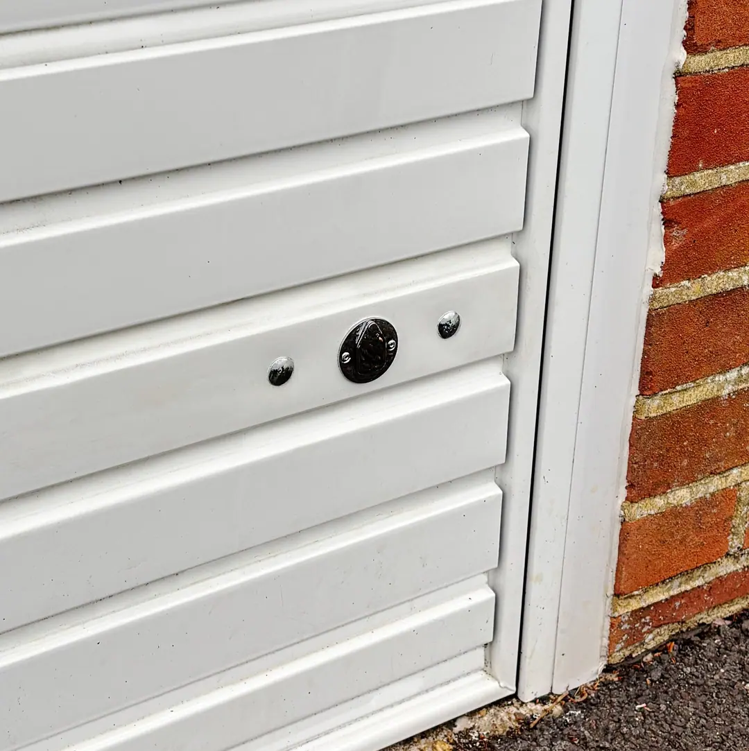The lower corner of a white panelled garage door beside a red brick pier, with a round chrome keyhole cover and two bolt heads
