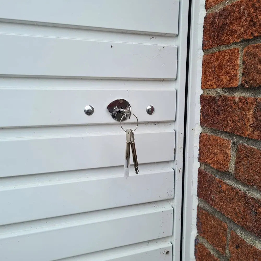 A white panelled garage door beside a red brick pier, with a chrome shoot bolt lock cover and keys hanging in the lock